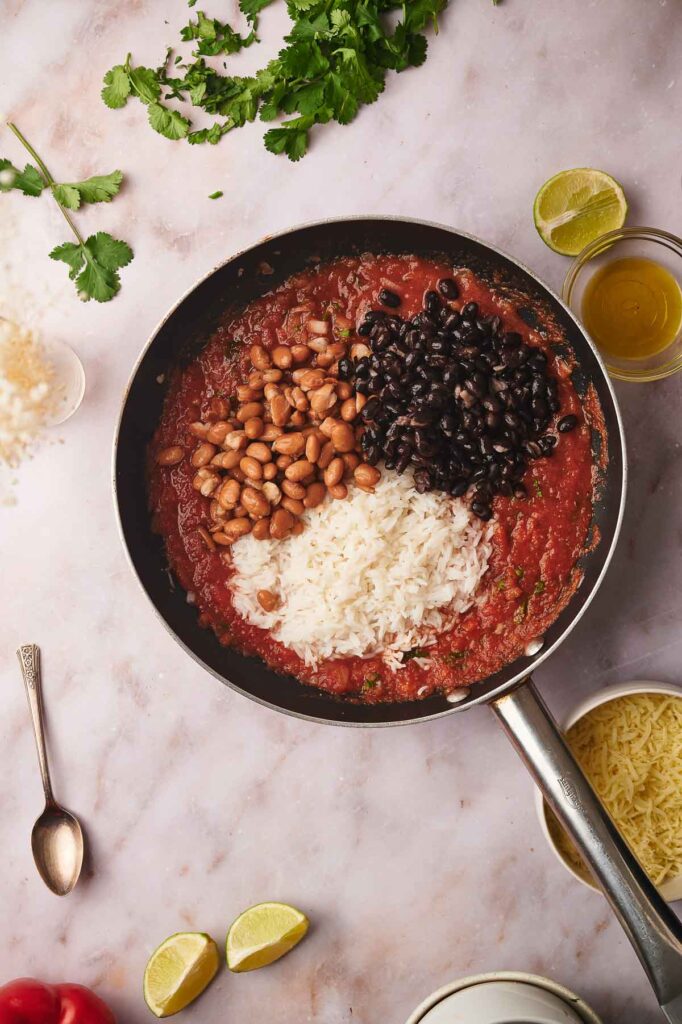 A skillet with tomato sauce, black beans, pinto beans, and white rice—reminiscent of the hearty filling in vegetarian stuffed peppers—sits on a marble surface, surrounded by cilantro, cheese, lime wedges, and a spoon.
