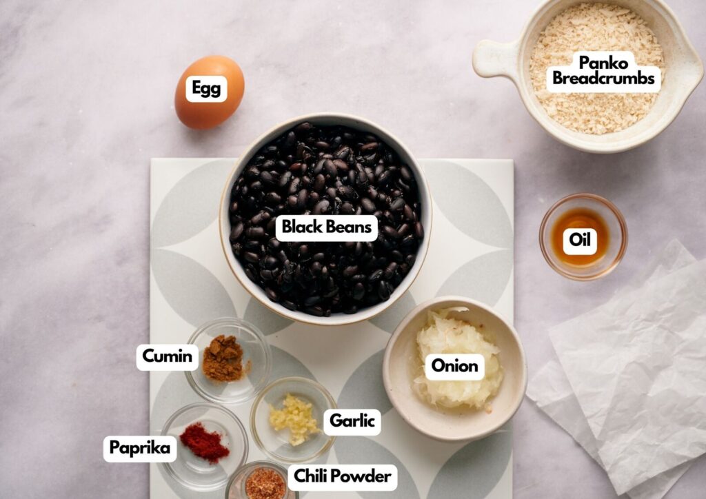 A flat lay shows labeled ingredients for a homemade black bean burger: black beans, egg, panko breadcrumbs, oil, onion, garlic, cumin, paprika, and chili powder arranged on a light surface.