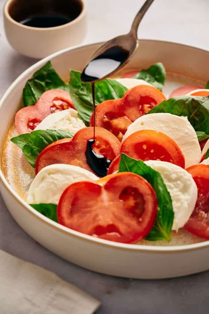 A spoon drizzling balsamic glaze over tomatoes, mozzarella slices, and basil leaves.