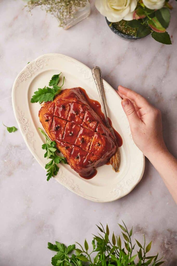 Cooked vegan ham on a serving dish, with a serving knife and a hand holding the dish.