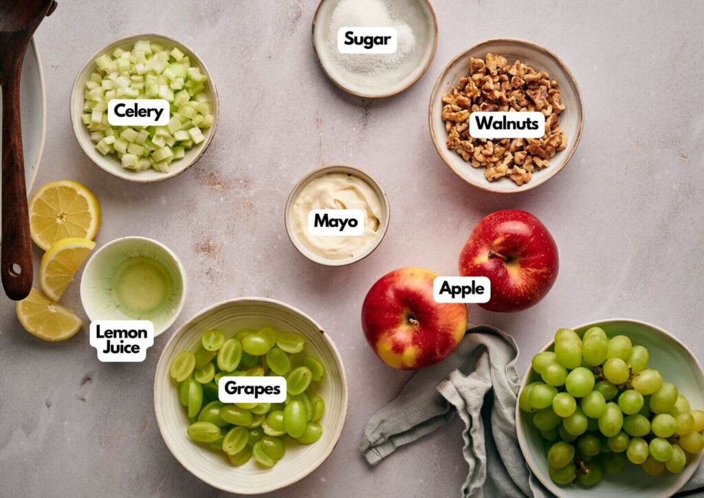 Bowls containing labeled ingredients for a classic Waldorf salad—grapes, apple, celery, walnuts, mayo, lemon juice, and sugar—are arranged on a light countertop.