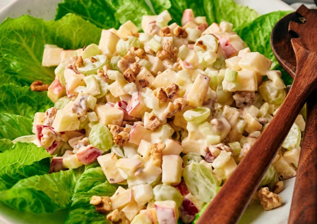 Enjoy a classic Waldorf salad featuring chopped apples, grapes, celery, walnuts, and a creamy dressing served on crisp romaine lettuce. Wooden salad servers are provided on the side for the perfect presentation of this refreshing waldorf salad.