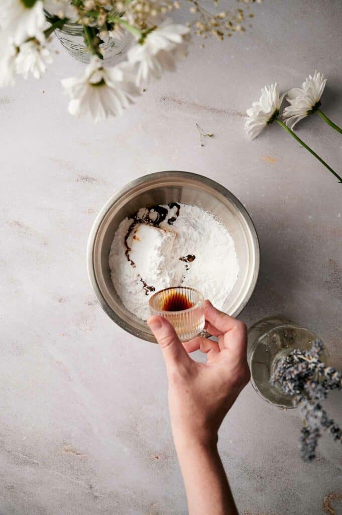 Confectioners sugar added to the bowl and someone pouring in vanilla extract.