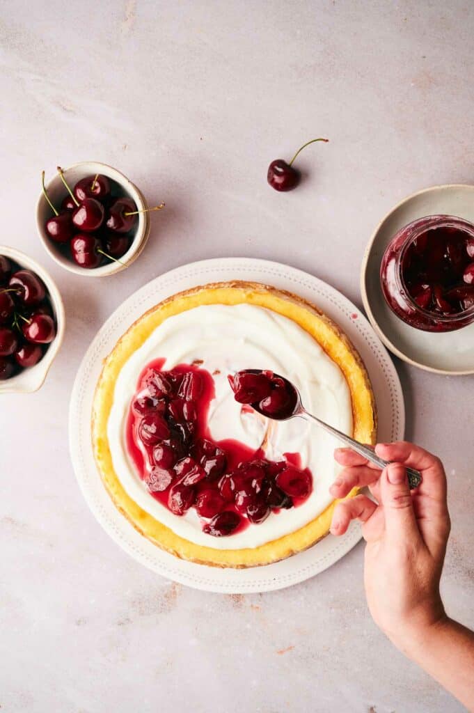 A person is spooning cherry sauce onto a cheesecake.