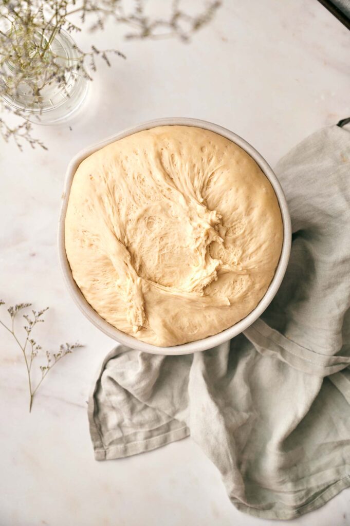 A bowl of risen bread dough sits on a light-colored surface next to a gray cloth and a vase with delicate white flowers, ready to be transformed into homemade french bread pizza.