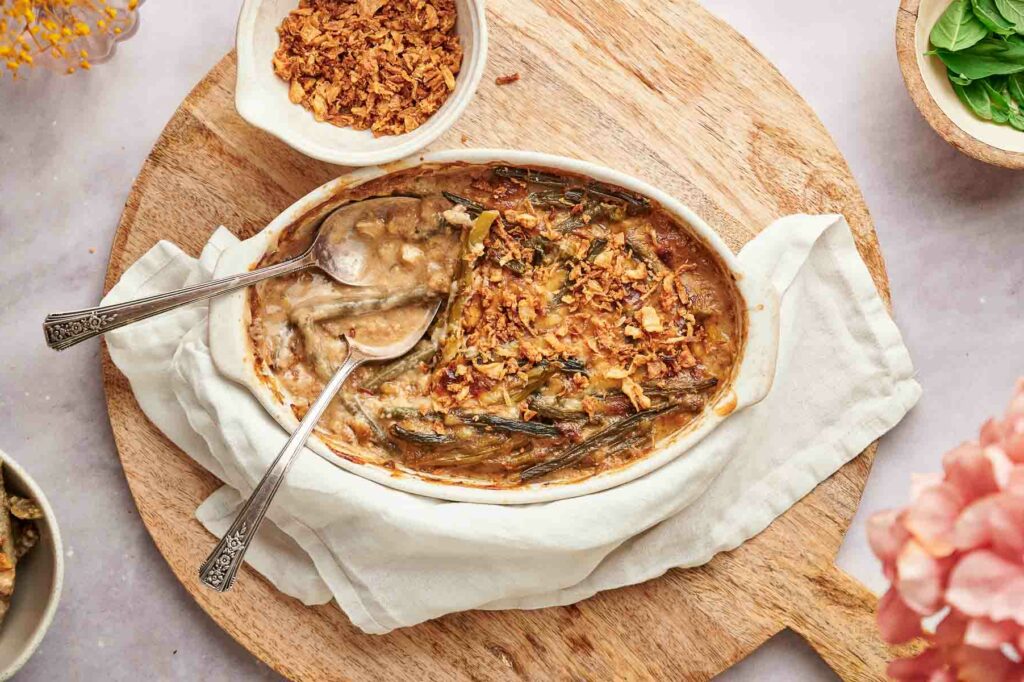 A dish filled with cheesy green bean casserole with a spoon on a wooden cutting board.