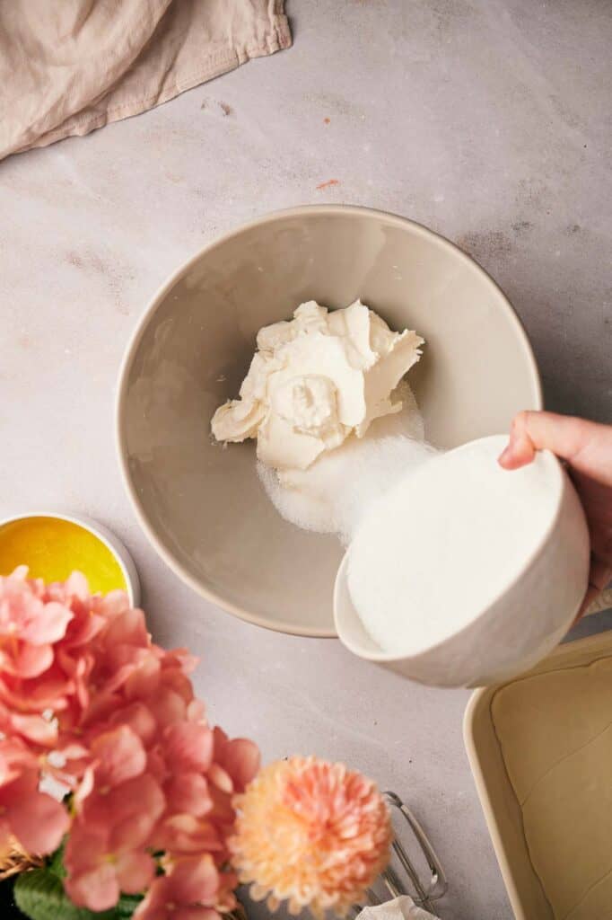 A person pouring sugar into a bowl of cream cheese.