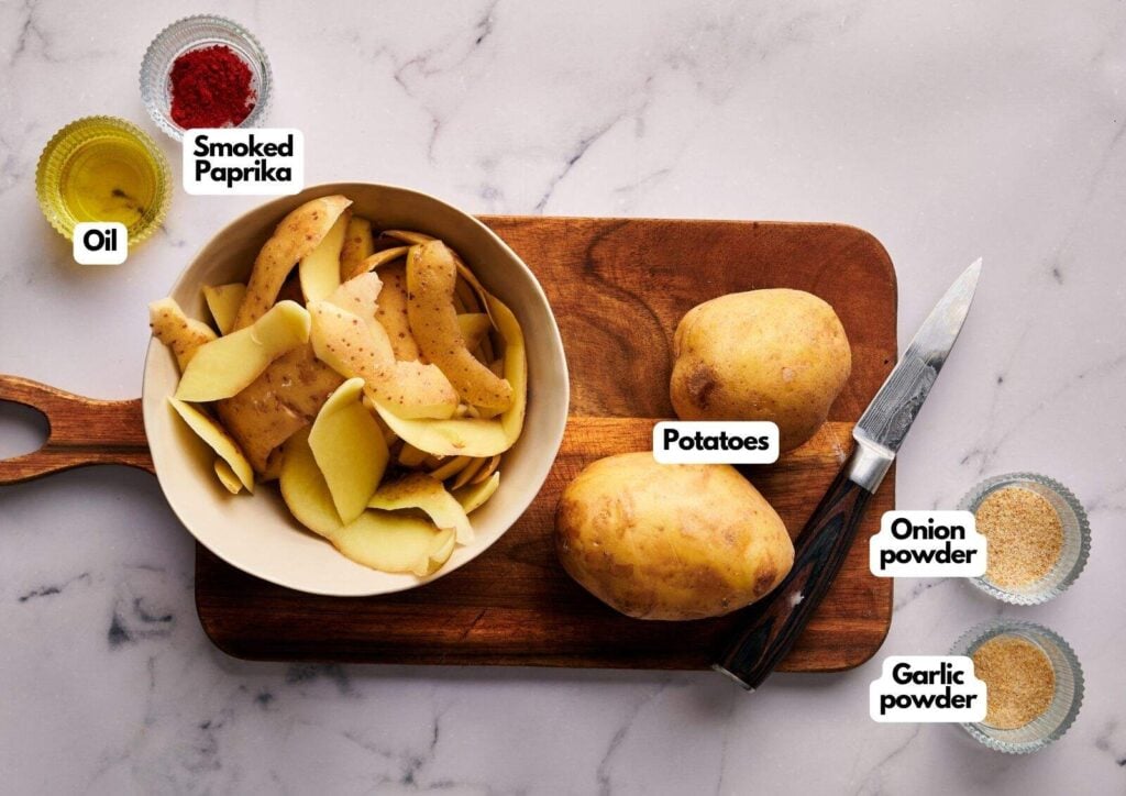 Sliced and whole potatoes on a cutting board with a knife, ready to be transformed into delicious breakfast potatoes, are surrounded by bowls labeled oil, smoked paprika, onion powder, and garlic powder on a marble surface.