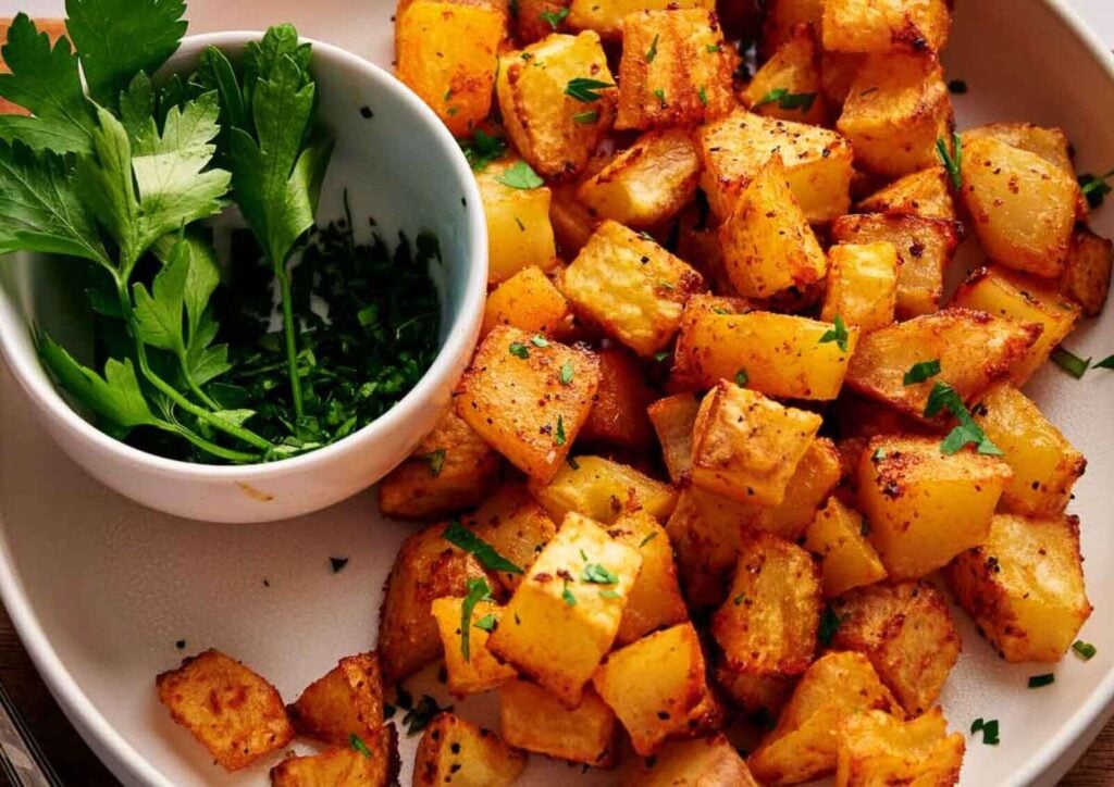 A plate of golden roasted breakfast potatoes cubes garnished with chopped herbs, served with a small bowl of fresh parsley.