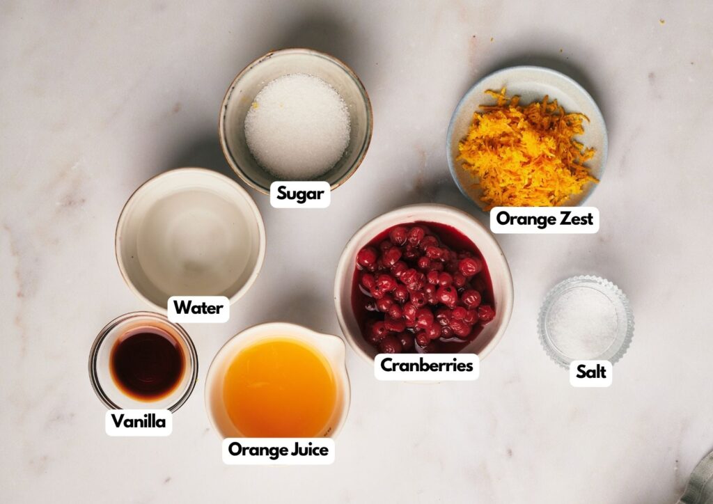 Six labeled bowls containing sugar, orange zest, water, cranberries, salt, vanilla, and orange juice—everything you need to make homemade cranberry sauce—are arranged on a white surface.