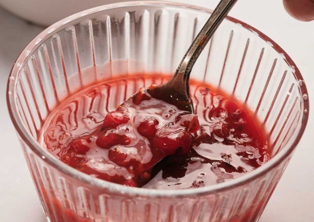 A glass container filled with chunky raspberry jam, with a fork partially submerged in the jam, reminiscent of the texture found in homemade cranberry sauce.