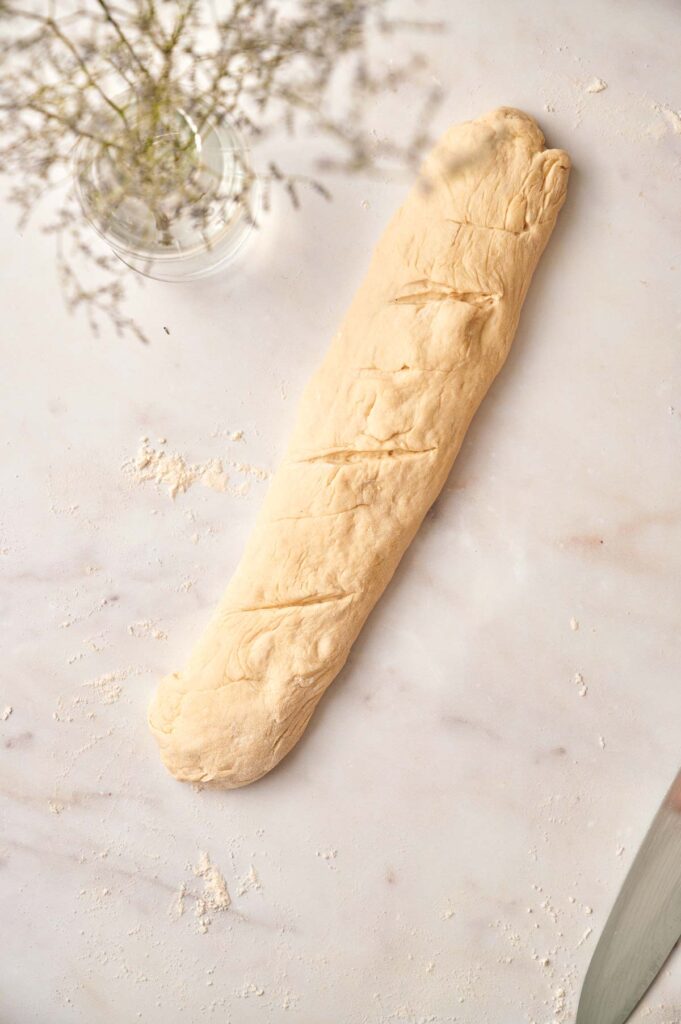 A long piece of french bread dough with slashes on top sits on a floured surface next to a glass vase with small flowers.