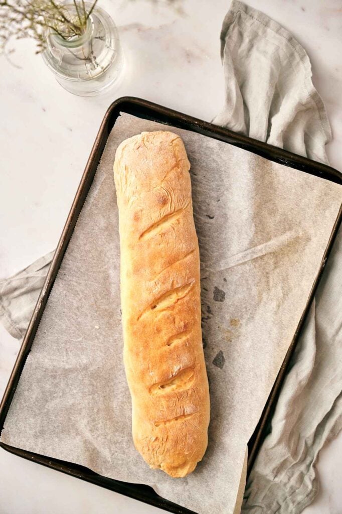 A freshly baked loaf of French bread rests on a parchment-lined baking sheet, placed on a marble surface with a light gray cloth and a vase of flowers nearby.