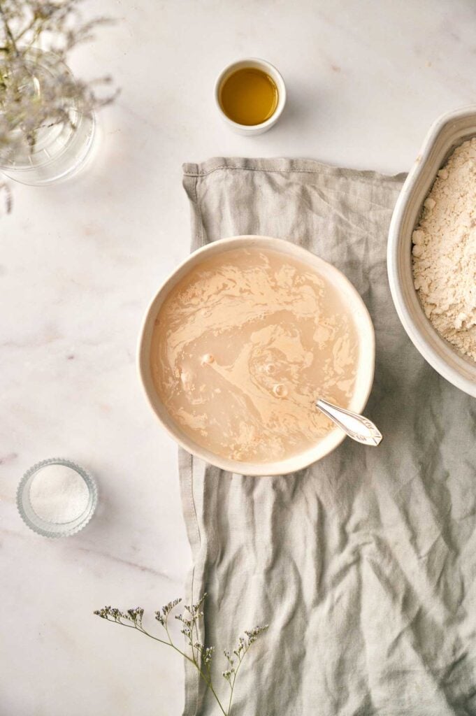 A bowl of yeast mixture with a spoon sits on a linen cloth, next to flour for french bread, a small cup of oil, and a dish of salt on a marble surface.