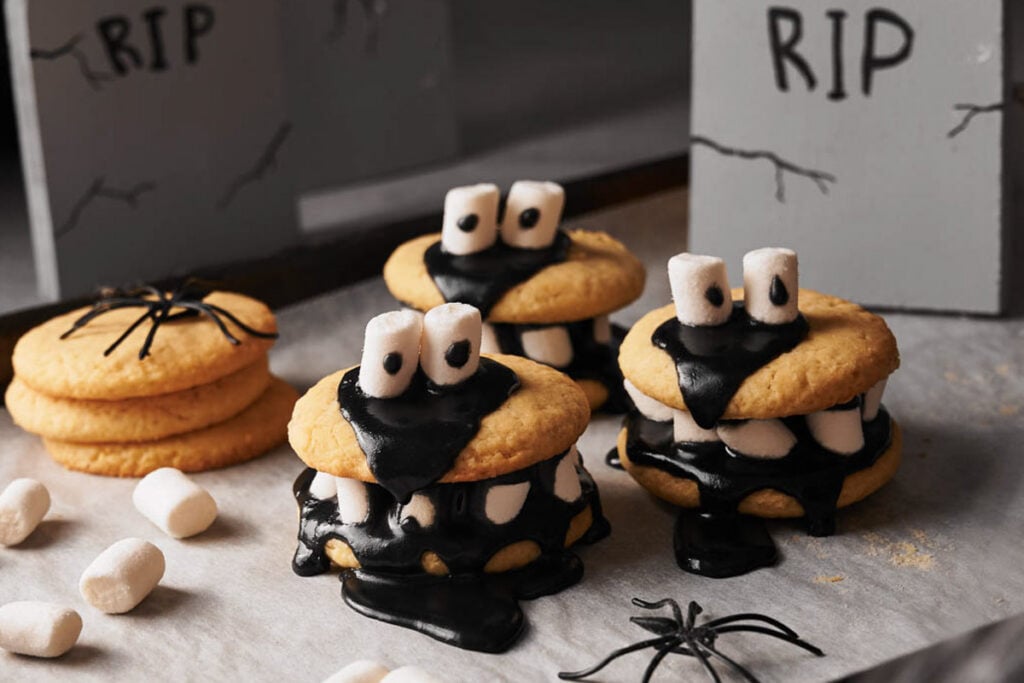 Three spooky Halloween cookies decorated with black icing and mini marshmallows for eyes and teeth, resembling monsters, sit on parchment next to plain cookies, marshmallows, and festive Halloween props.