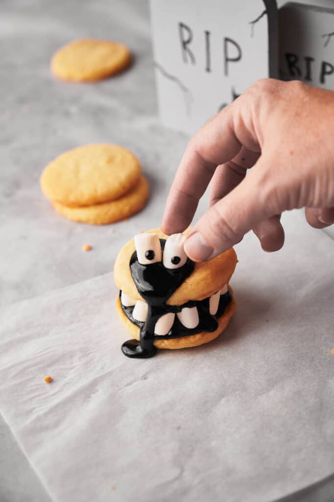 A person is placing marshmallow eyes on top of the cookie.