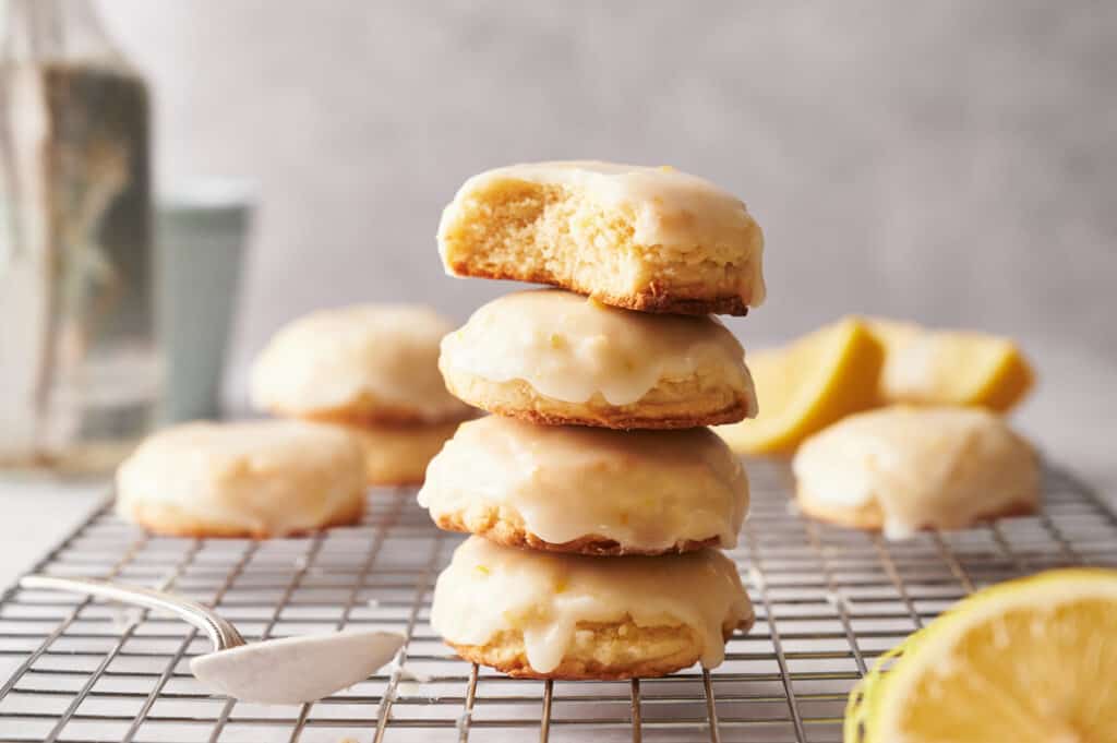 A stack of lemon cookies on a cooling rack with a lemon half and a spoon with lemon glaze on.
