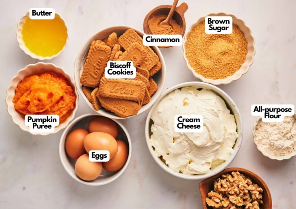 Assorted baking ingredients in bowls, including butter, pumpkin puree, eggs, Biscoff cookies, cinnamon, cream cheese, brown sugar, all-purpose flour, and walnuts&mdash;everything you need to create a delicious pumpkin cheesecake.