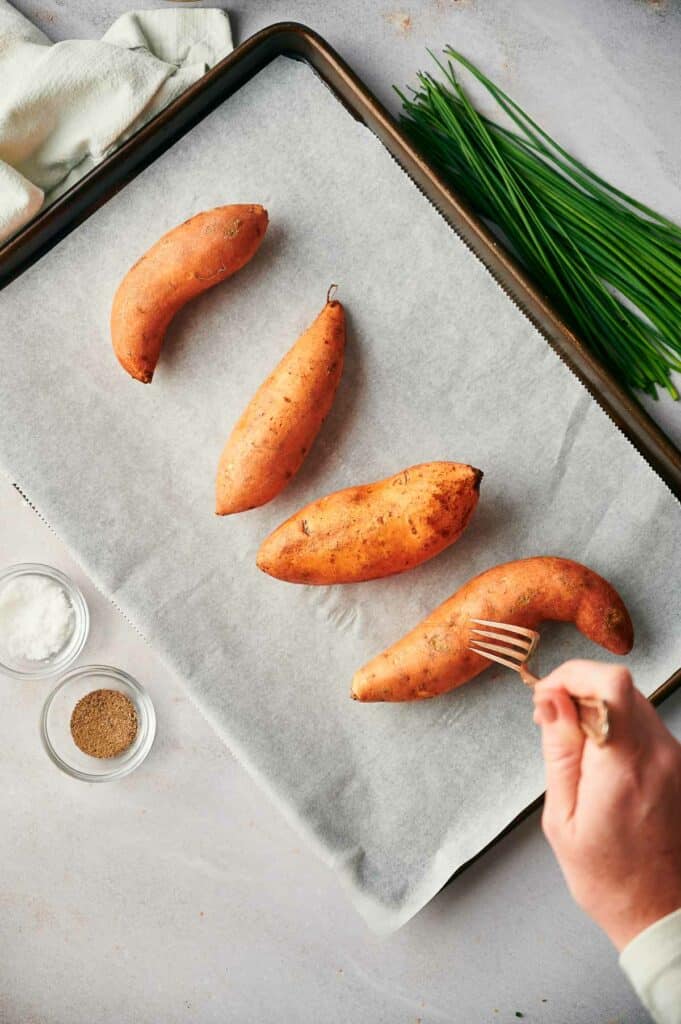 A person pricking sweet potatoes with a fork on a baking sheet.