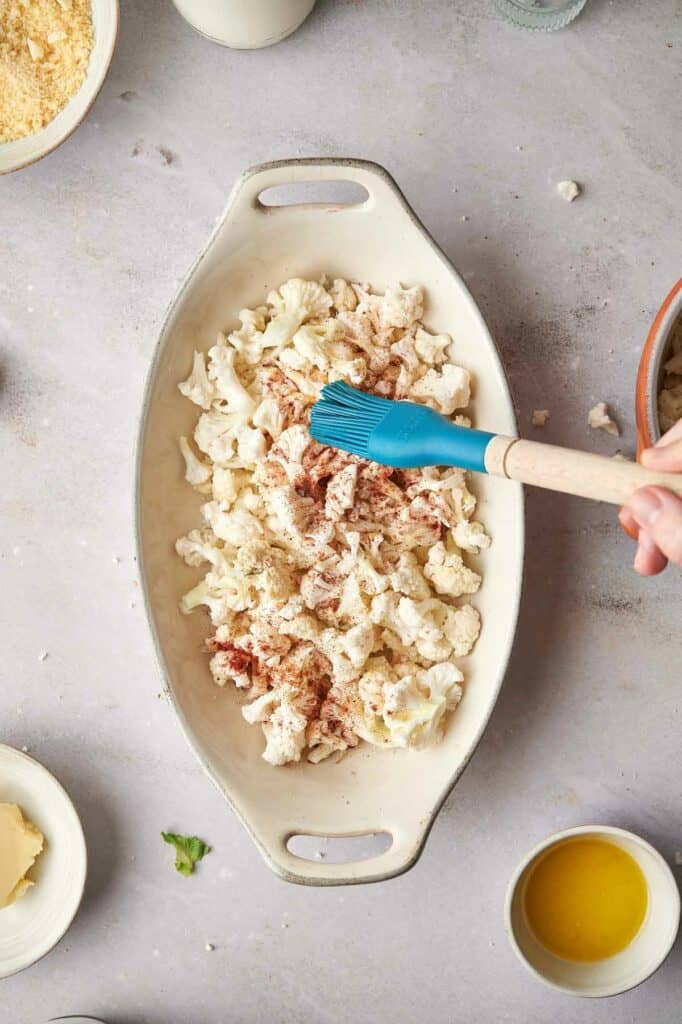 A person using a blue spoon to coat cauliflower in a bowl.
