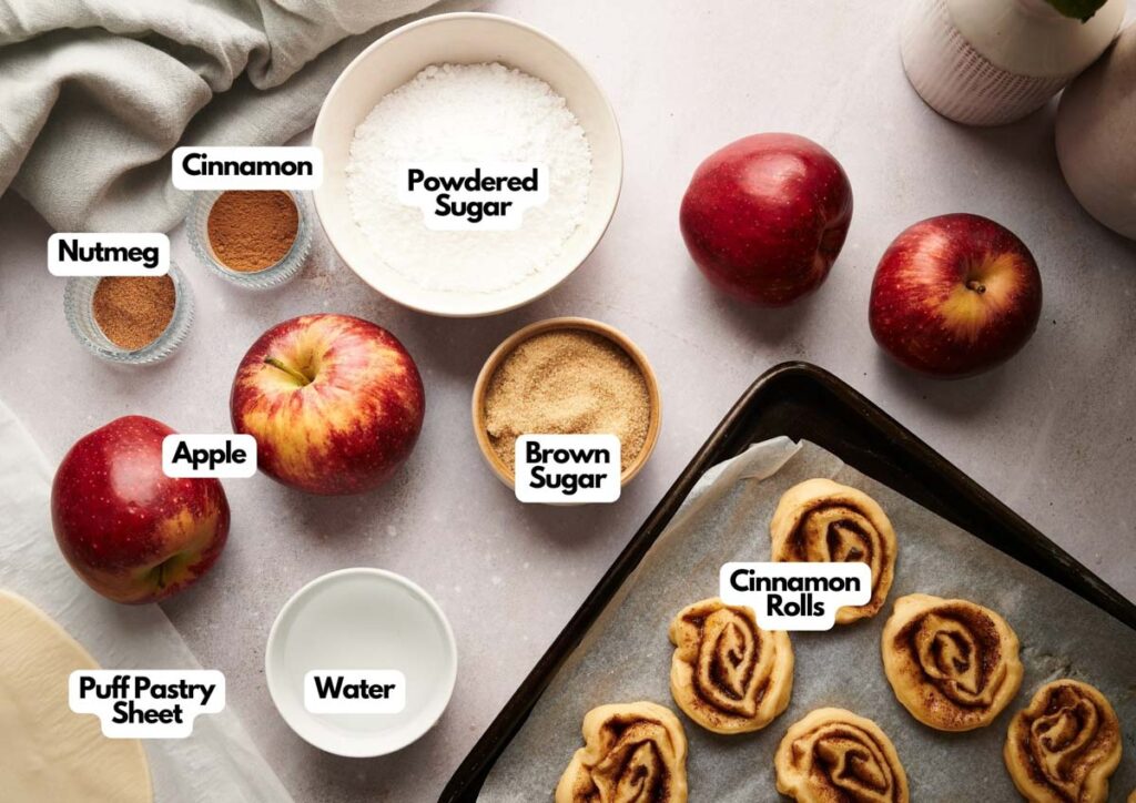 Ingredients for cinnamon apple rolls, inspired by a cinnamon roll apple pie, including apples, cinnamon, nutmeg, powdered sugar, brown sugar, water, and puff pastry sheet arranged on a countertop with baked rolls on a tray.