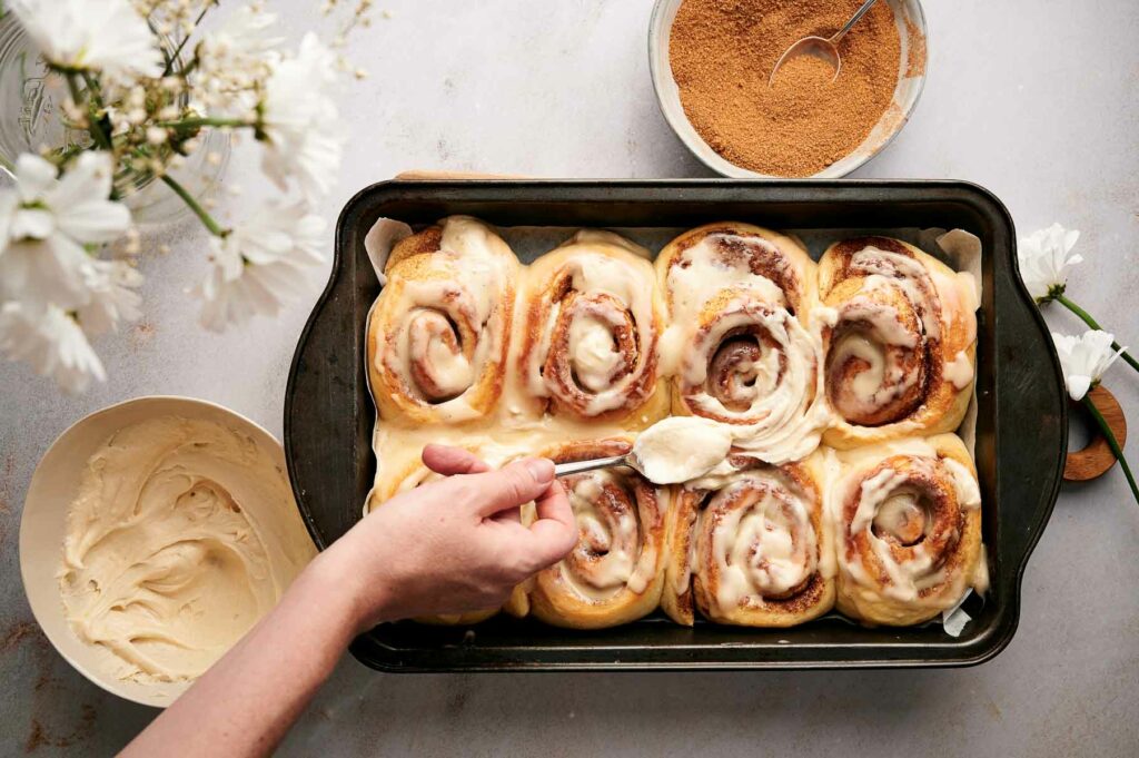 Cinnamon rolls in a pan with someone putting icing on top.