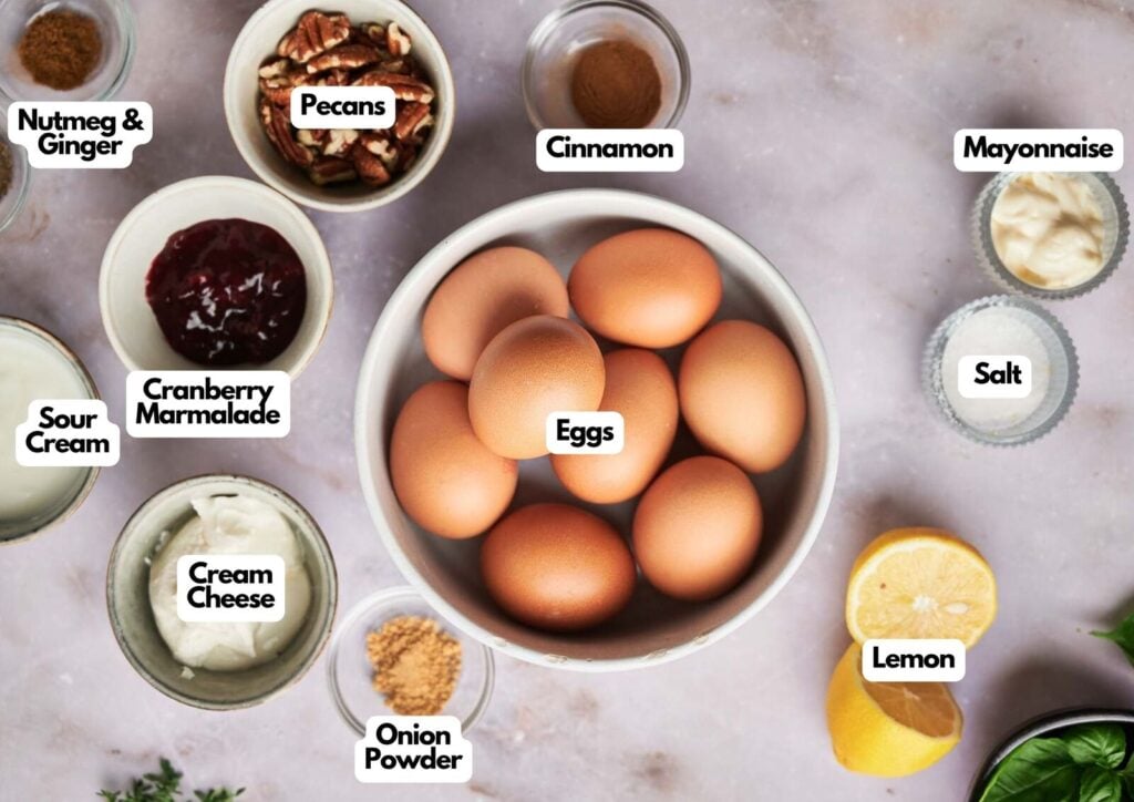 A bowl of eggs for deviled eggs is surrounded by labeled ingredients including mayonnaise, salt, lemon, cream cheese, sour cream, cranberry marmalade, nutmeg & ginger, pecans, cinnamon, and onion powder.
