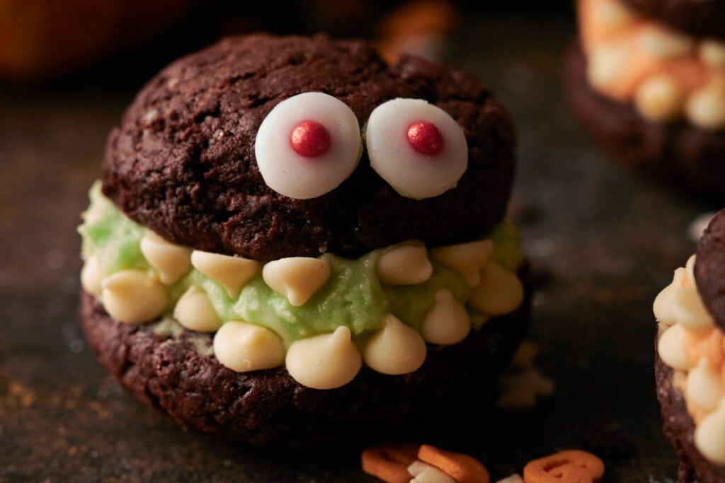 A Whoopie Pie Halloween Monster, this chocolate cookie sandwich is decorated with candy eyes, green frosting, and white chocolate chips to create a fun and spooky monster face.