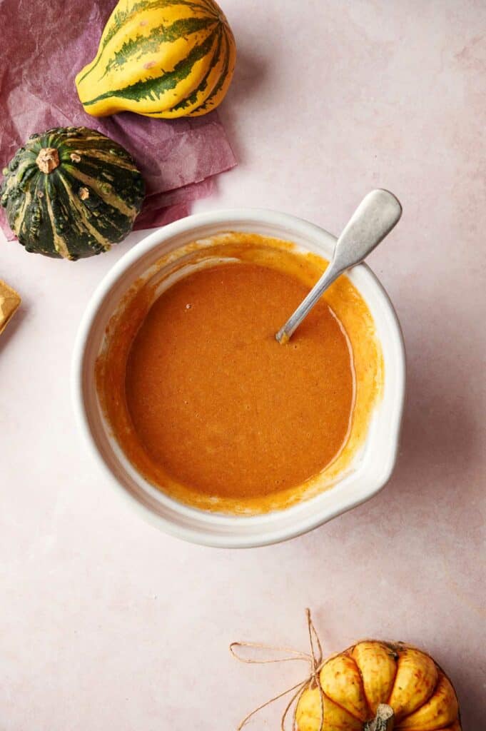 A bowl of pumpkin pie filling with a spoon and pumpkins.