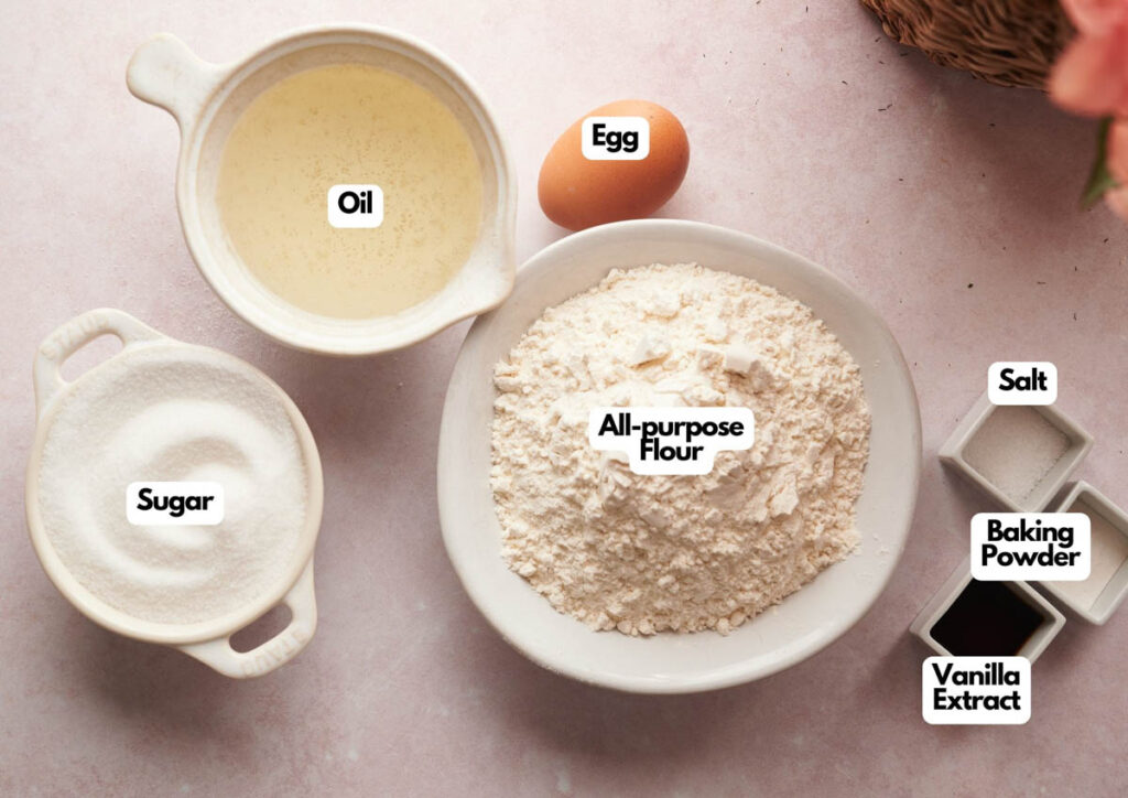 Bowls containing oil, sugar, all-purpose flour, salt, baking powder, and vanilla extract, plus a single egg, are arranged on a light pink surface&mdash;perfect ingredients for making Whoopie Pie Halloween Monsters.