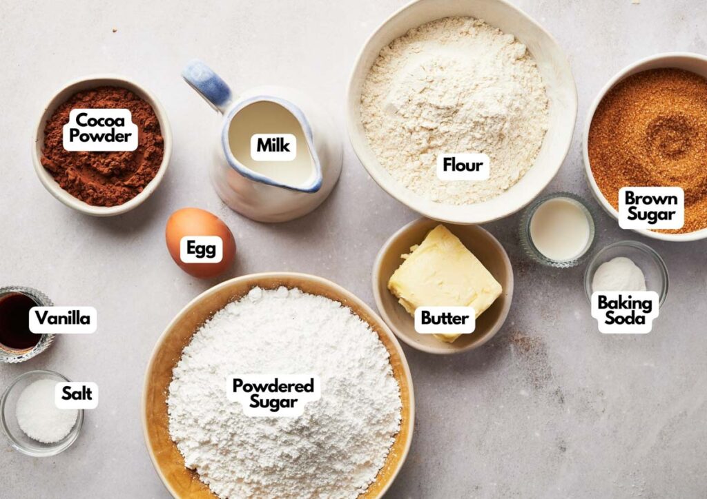 Bowls and containers of cocoa powder, milk, flour, brown sugar, vanilla, salt, powdered sugar, butter, an egg, and baking soda&mdash;perfect for making whoopie pie&mdash;are arranged on a countertop with labels identifying each ingredient.