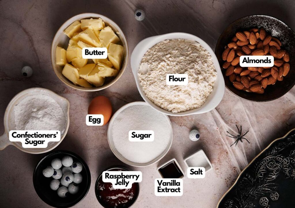 Assorted baking ingredients in bowls labeled as butter, flour, almonds, confectioners&rsquo; sugar, egg, sugar, raspberry jelly, vanilla extract, and salt on a brown surface&mdash;perfect for making spooky Witch Finger Cookies.