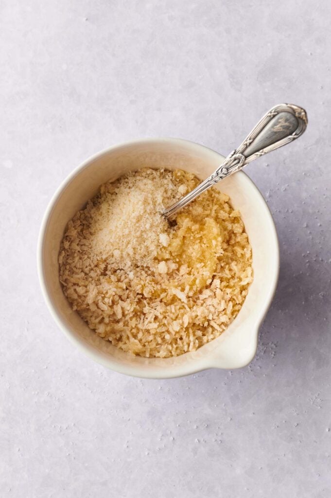 A white bowl containing dry breadcrumbs, grated parmesan cheese, and a metal spoon, prepared as key ingredients for a delicious zucchini casserole, placed on a light textured surface.