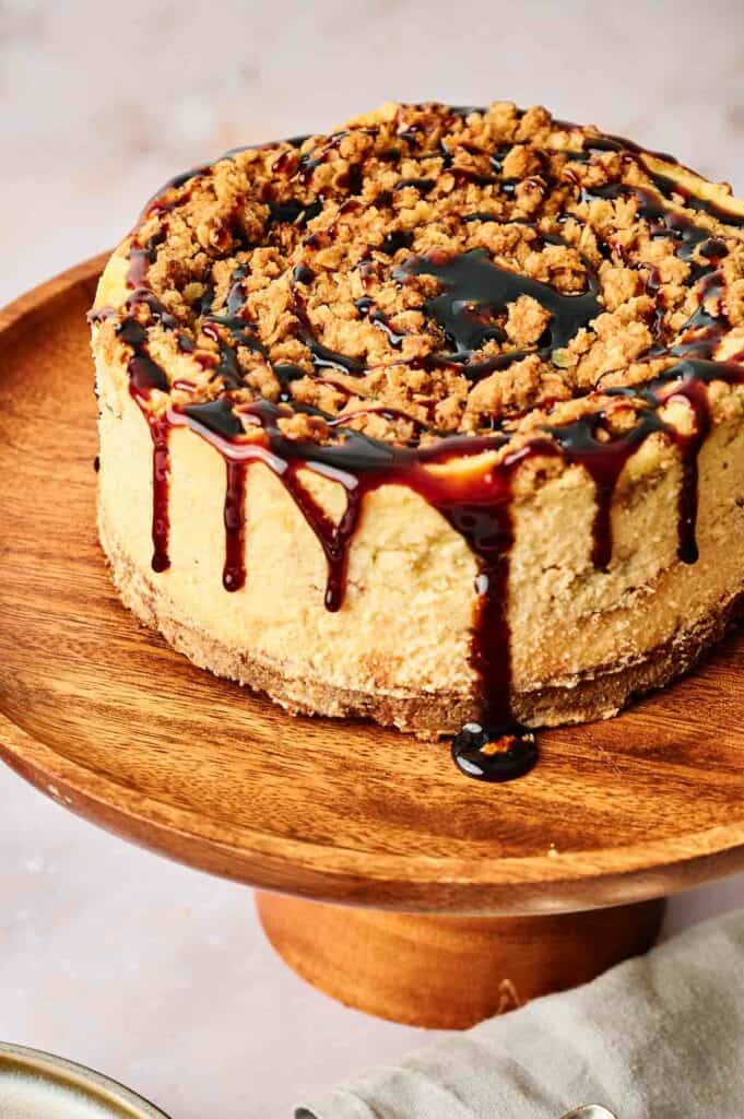 An apple pie stuffed cheesecake is sitting on top of a wooden cake stand.