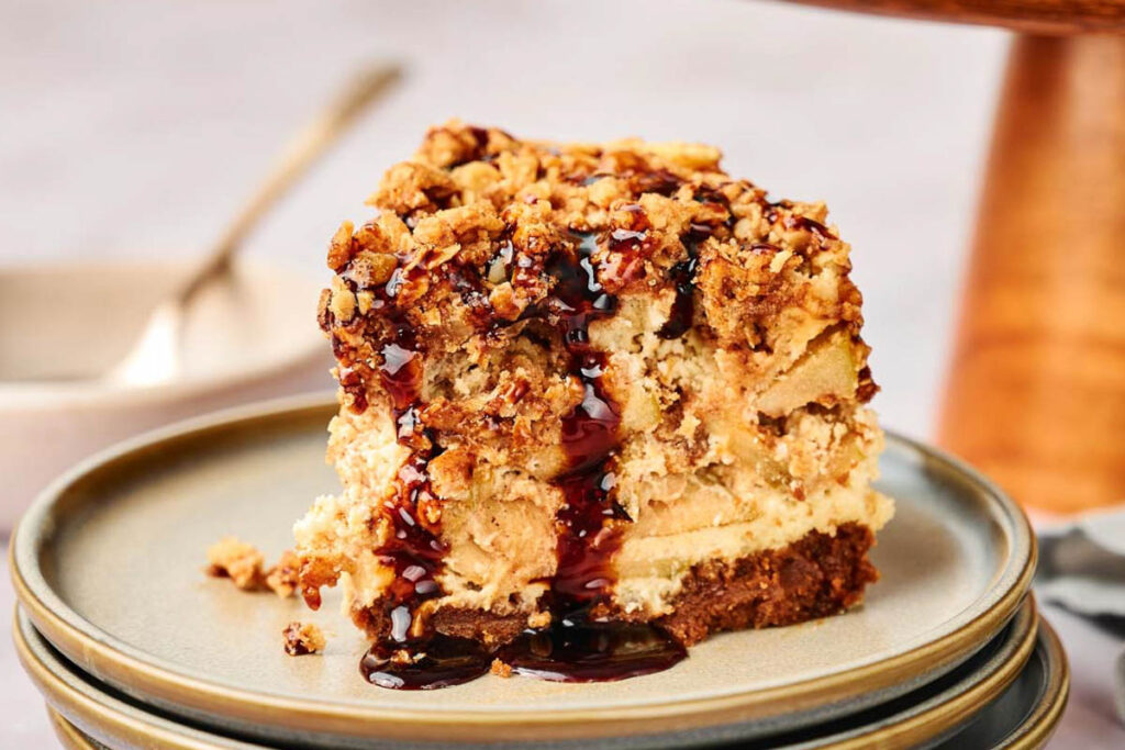 A slice of crumb-topped apple pie stuffed cheesecake with chocolate sauce drizzled on top, served on a stack of ceramic plates.