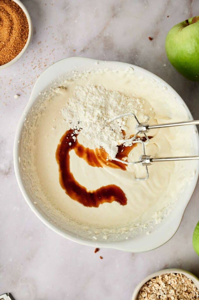 A bowl with whipped cream and vanilla extract, with apples and a spoon next to it.