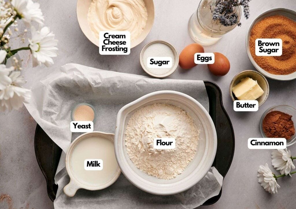Baking ingredients arranged on a table include flour, milk, yeast, eggs, sugar, brown sugar, butter, cinnamon—perfect for making cinnamon rolls—and a bowl of cream cheese frosting.
