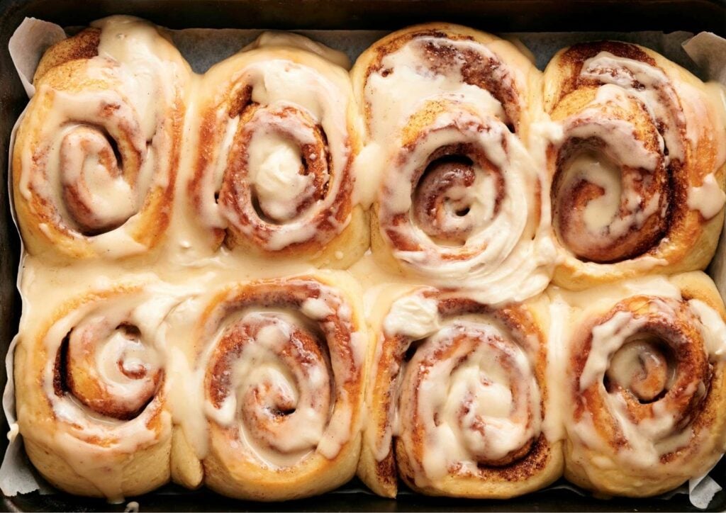 Eight soft cinnamon rolls in a baking tray, topped with melted icing and arranged closely together.