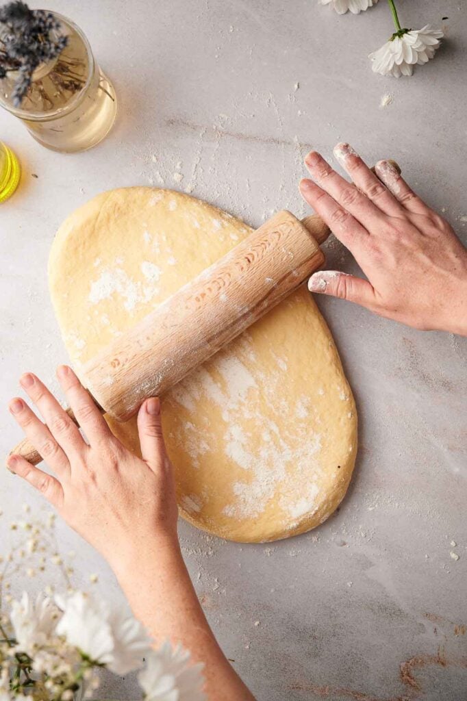A person rolls out dough for cinnamon rolls with a wooden rolling pin on a floured surface, with white flowers and a drink visible nearby.