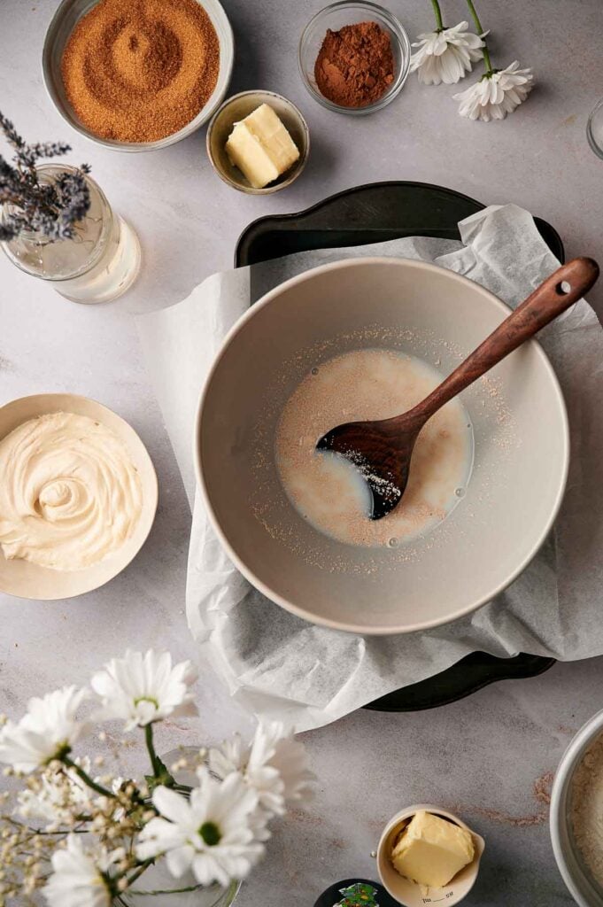 A mixing bowl with liquid and a wooden spoon sits on parchment paper, surrounded by bowls of butter, cocoa, sugar, and frosting—perfect ingredients for making homemade cinnamon rolls—with flowers nearby.