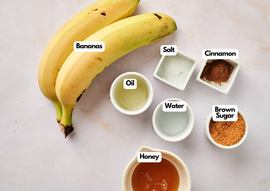 Two bananas, small bowls of oil, honey, water, brown sugar, salt, and cinnamon are arranged on a light surface with each item labeled&mdash;perfect for creating apple pie stuffed cheesecake-inspired treats.