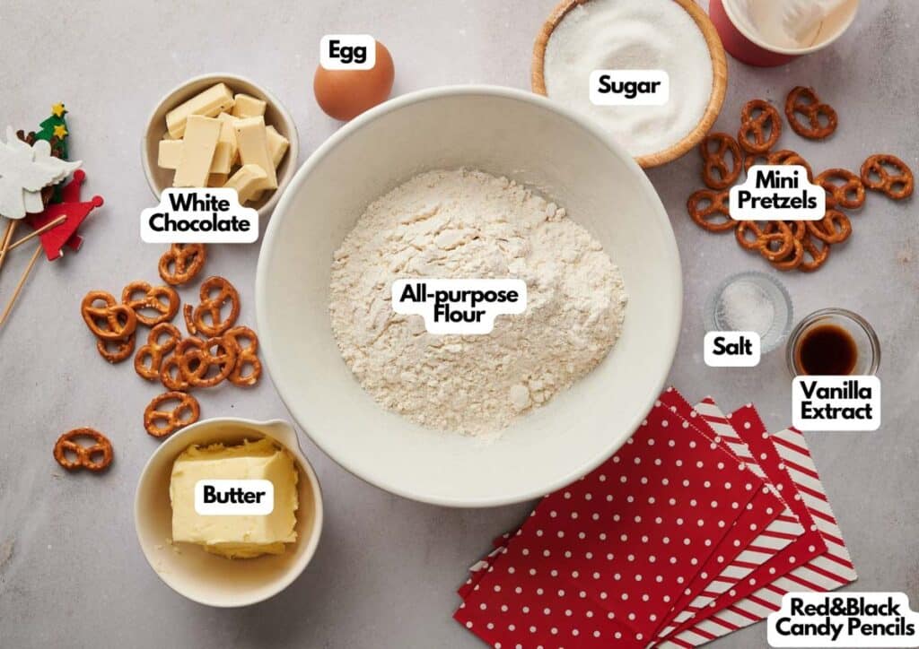 Baking ingredients for reindeer Christmas cookies, including all-purpose flour, white chocolate, butter, egg, sugar, mini pretzels, salt, vanilla extract, and red & black candy pencils arranged on a countertop.