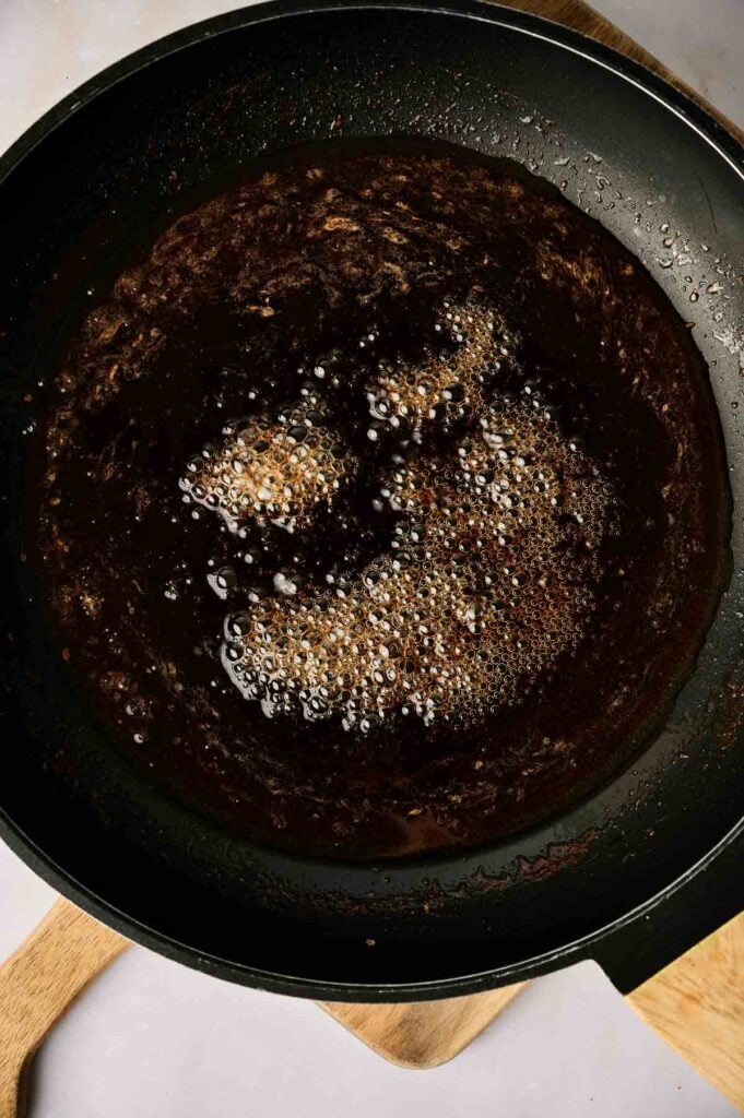 A close-up of a black skillet with a bubbling dark brown sauce or liquid, possibly caramelizing for fried bananas, cooking on a wooden board.
