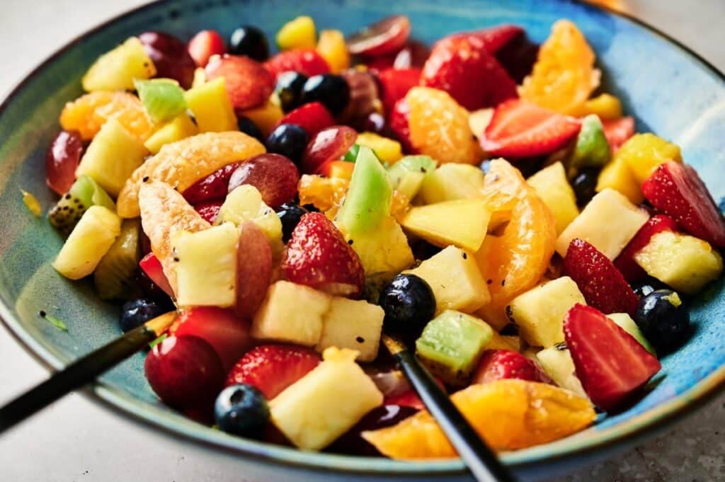 A bowl filled with a mixed fruit salad including strawberries, blueberries, kiwi, pineapple, grapes, and orange slices, alongside two black utensils—perfect as a fresh pairing with air fryer chocolate crinkle cookies.