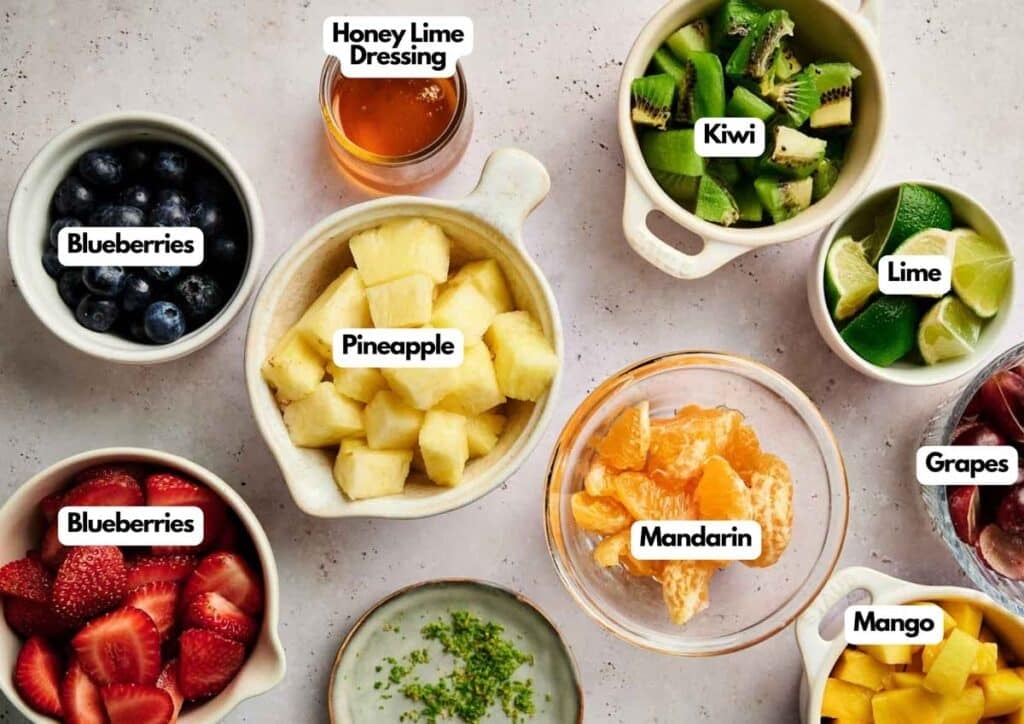 Bowls of assorted fruits—blueberries, strawberries, pineapple, mandarin, grapes, kiwi, mango, lime wedges—and a cup of honey lime dressing sit on a light surface beside Air Fryer chocolate crinkle cookies.