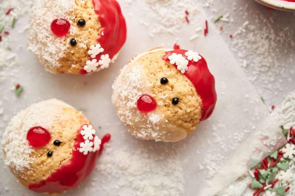 Three cupcakes decorated as Santa Claus faces, with red icing hats, white coconut beards, candy eyes, and red candy noses, are arranged on a lightly dusted surface beside air fryer chocolate crinkle cookies.
