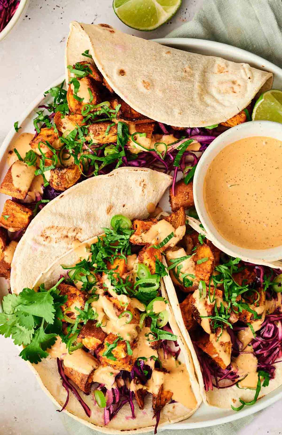 Sweet Potato Tacos filled with roasted sweet potatoes, shredded purple cabbage, chopped cilantro, and a creamy chipotle sauce, served on a plate with lime wedges and a small bowl of extra sauce.