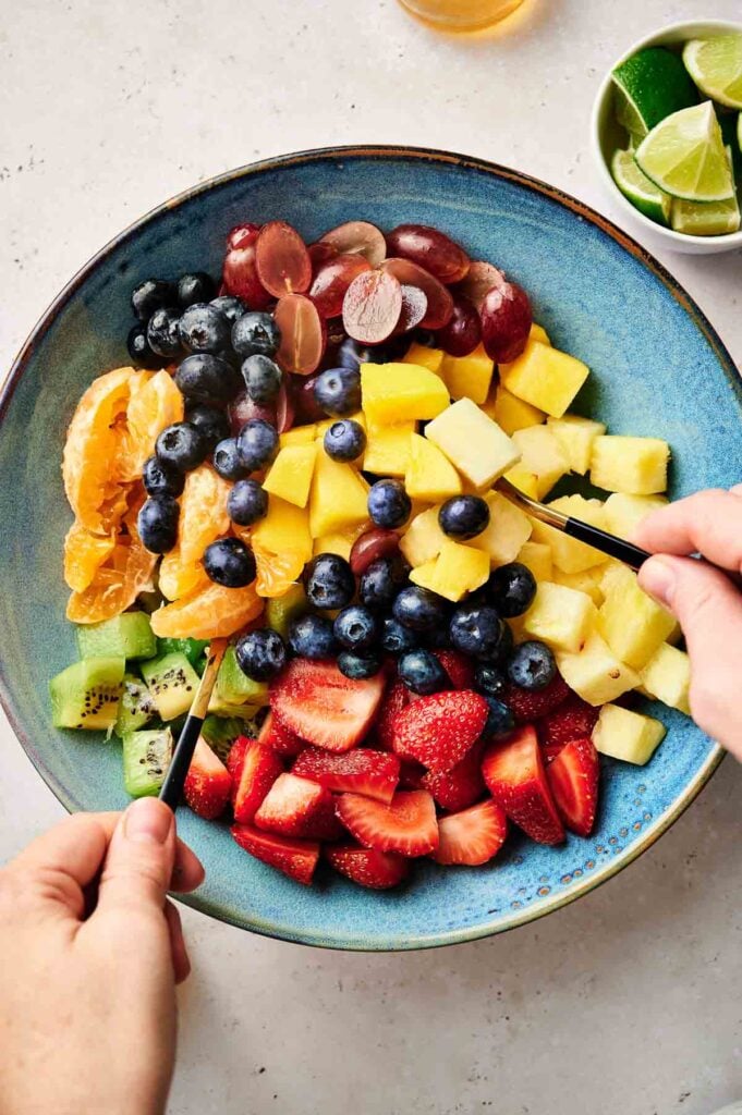 A bowl of healthy fruit salad featuring assorted fresh fruit including strawberries, kiwi, blueberries, grapes, pineapple, and orange segments, with hands mixing the vibrant salad using two utensils.