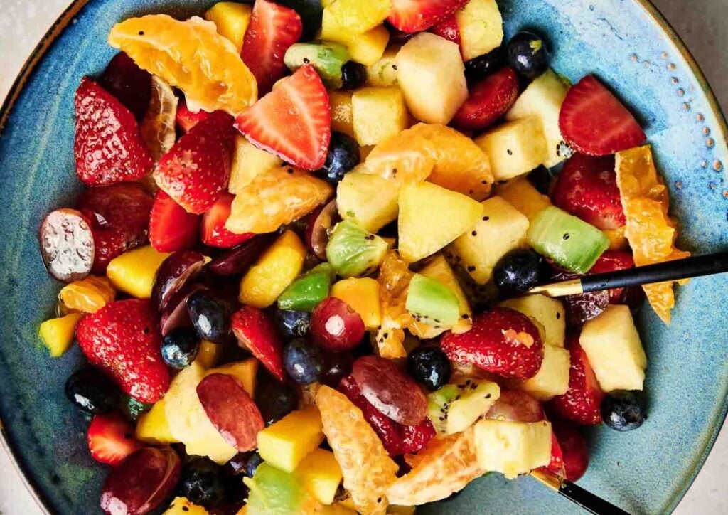 A blue bowl filled with a colorful mix of sliced strawberries, grapes, blueberries, kiwi, orange segments, and melon cubes creates a vibrant healthy fruit salad, with a pair of black chopsticks on the side.