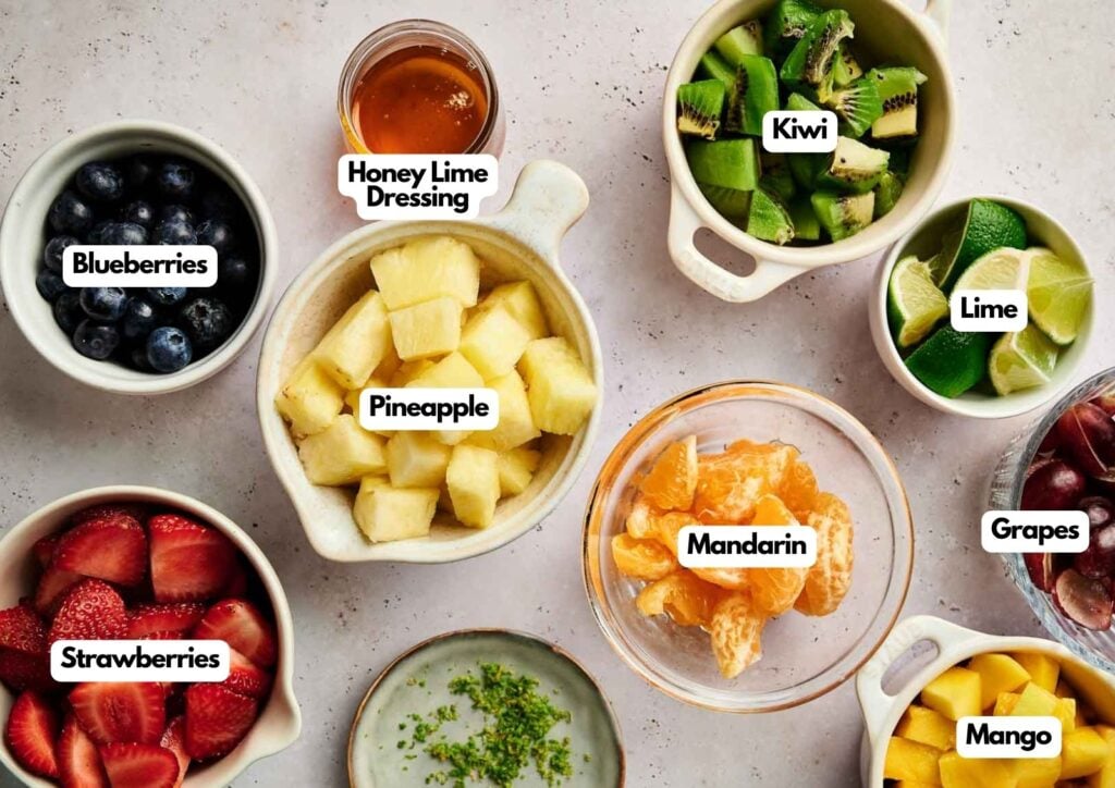 Overhead shot of fruit salad ingredients including blueberries, sliced strawberries, pineapple chunks, kiwi, mandarin segments, grapes, and mango cubes with honey lime dressing.